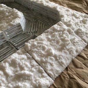 May include: Close-up of a set of white textured tiles, resembling snow, arranged on a brown paper surface. The tiles are used for creating a miniature winter scene.