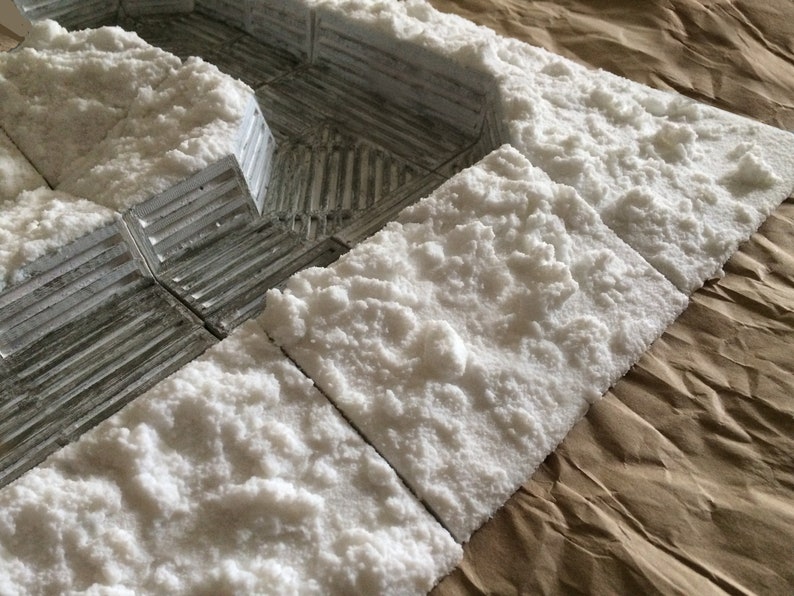 May include: Close-up of a set of white textured tiles, resembling snow, arranged on a brown paper surface. The tiles are used for creating a miniature winter scene.