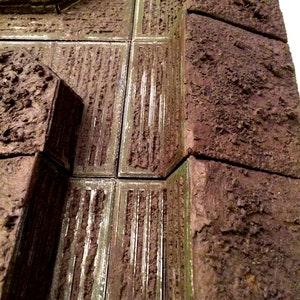 Pode incluir: Um close-up de um modelo texturizado de um edif&iacute;cio marrom e verde com uma abertura retangular. O modelo &eacute; feito de um material que se assemelha a pedra ou concreto.