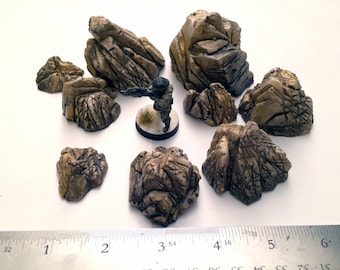May include: A set of nine brown, textured, rocky terrain pieces for tabletop gaming. The rocks are various sizes and shapes, with some having a more jagged appearance than others.