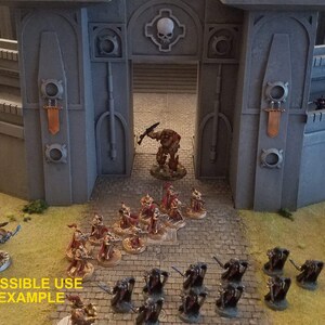 May include: A miniature wargaming scene featuring a fortress gate with a skull emblem. Several groups of painted miniature figures are positioned on a textured surface, suggesting a battle scenario. The text "POSSIBLE USE EXAMPLE" is visible.
