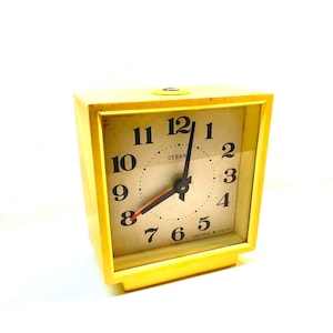 Pode incluir: Um despertador quadrado amarelo vintage com um mostrador creme. O mostrador apresenta algarismos pretos, ponteiros pretos e um ponteiro de segundos vermelho. A palavra "CEBAH" está impressa no mostrador. O relógio está sobre um fundo branco.