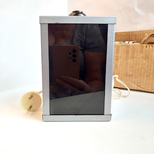 Puede incluir: Un espejo rectangular con marco plateado y superficie reflectante negra. El espejo tiene un pequeño botón negro redondo en la parte superior y un enchufe eléctrico blanco sujeto a un cable. Una caja de cartón marrón está en el fondo.