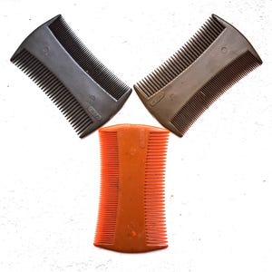 Op de afbeelding: Drie plastic luizenkammen in grijstinten, bruin en oranje zijn tegen een witte achtergrond geplaatst. Elke kam heeft fijne tanden en een gebogen handvat. De kammen worden waarschijnlijk gebruikt om luizen en neten uit het haar te verwijderen.