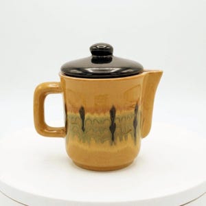 May include: A ceramic teapot with a glossy mustard yellow body featuring a decorative drip glaze pattern in shades of green and brown. The teapot has a square-shaped handle, a short spout, and a removable lid with a dark brown, glossy finish.