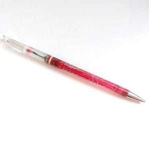 May include: A clear pen with a red floral design encased within the barrel. The pen has a silver tip and a decorative band near the top. The pen is angled on a white surface.