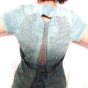 Puede incluir: Camiseta azul verdosa de manga corta con un aspecto desgastado. La espalda presenta un diseño recortado con un detalle trenzado y alas de ángel con pedrería. La parte inferior de la camiseta tiene un diseño rasgado.