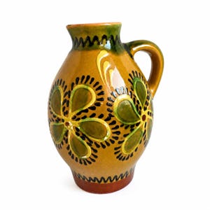 May include: A glazed ceramic pitcher with a mustard yellow base colour, featuring hand-painted green floral motifs outlined in black. The vessel has a rounded body, a single curved handle, and a dark brown base with a zigzag pattern at the rim.