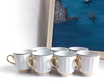 May include: Six white porcelain teacups with gold handles and trim. Each cup has a black line around the rim and a gold band at the base. The cups are arranged in a row. A painting of a seascape is in the background.