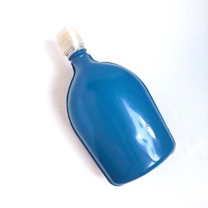 May include: A blue hot water bottle with a white and clear plastic stopper. The bottle has a rounded shape and is made of a shiny, smooth material. The stopper is attached to the bottle's neck.