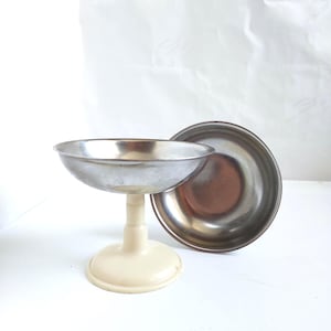 May include: A vintage metal pedestal bowl with a cream-colored base and stem, next to a matching metal bowl. The bowl has a silver finish and a rounded shape, perfect for serving desserts or displaying small items.