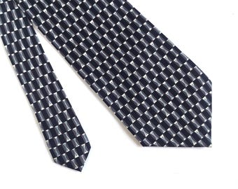 Puede incluir: Una corbata azul marino con un patrón geométrico repetitivo de pequeños cuadrados conectados en negro, gris y blanco. La corbata está en ángulo sobre una superficie blanca, mostrando su diseño y textura.