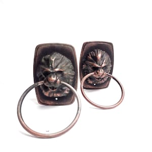 May include: Two antique-style door knockers with lion head designs. Each knocker is made of metal and has a ring attached. The knockers are a dark brown color with a copper finish.