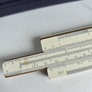 Può includere: Un regolo calcolatore vintage con segni bianchi e marroni. Il regolo ha una sezione centrale scorrevole ed è utilizzato per calcoli matematici. I segni includono numeri e scale per vari calcoli. È uno strumento classico.