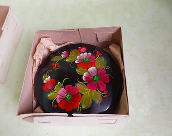 May include: A round black lacquer box featuring a hand-painted floral design with vibrant red, pink, and white petals accented by green leaves and small red berries. The box is nestled inside a light tan cardboard container with tissue paper.