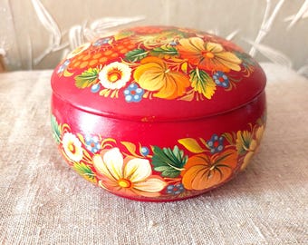 May include: A round, red wooden box with a lid, hand-painted with a floral design. The flowers are yellow, orange, and white, with green leaves and blue accents. The box is sitting on a beige linen surface.