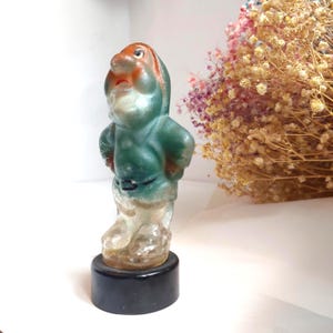 May include: A vintage figurine of a dwarf character, featuring a green hooded jacket, orange face, and white beard. The figurine is mounted on a black base. The dwarf is standing with hands on hips. The figurine is likely made of glass or a similar translucent material.