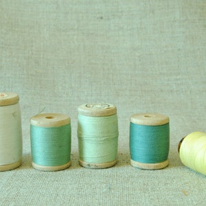 May include: Five spools of thread, four are wooden with green thread and one is wooden with white thread. A yellow spool of thread is lying on its side.