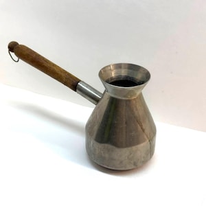Puede incluir: Una cafetera turca de color plateado, también conocida como cezve, con un mango de madera. La cafetera tiene una base ancha que se estrecha hacia un cuello estrecho y un borde acampanado. El mango está sujeto con una banda de metal.