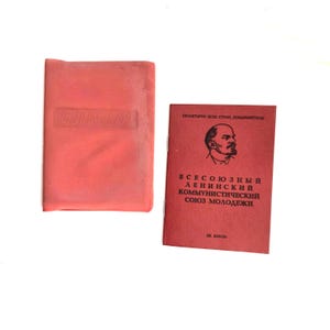 May include: Two vintage red items on a white background. One is a rectangular case with the embossed letters "ВЛКСМ". The other is a red booklet with a portrait and Cyrillic text. The booklet is titled "Всесоюзный Ленинский Коммунистический Союз Молодежи".