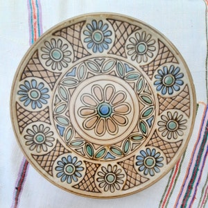 Könnte beinhalten: Ein dekorativer Keramikteller mit floralem Design. Der Teller hat einen beigefarbenen Hintergrund mit blauen, grünen und braunen Blumenmustern. Die Blumen sind kreisförmig angeordnet. Der Teller hat einen erhöhten Rand.