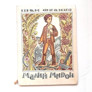 Puede incluir: Portada de libro vintage con un joven en traje marrón sosteniendo una llama, rodeado de ilustraciones florales y agrícolas. El título y el nombre del autor están en una fuente estilizada.