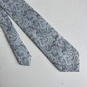 May include: A light blue necktie with a paisley pattern in shades of blue and gray. The tie is made of a textured fabric and is displayed on a white surface. The tie is a classic accessory.