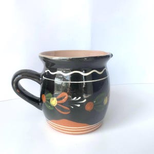 May include: A black ceramic jug with a rounded body and a single handle. The exterior features a hand-painted floral motif in orange, green, and yellow, accented by a white wavy line near the rim and three horizontal white lines at the base.