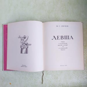 May include: An open book with a black and white illustration of a blacksmith's anvil and hammer on the left page. The right page has the title "Levsha" in large black letters and the text "A tale of the Tula Lefty and the Steel Flea" in smaller black letters.