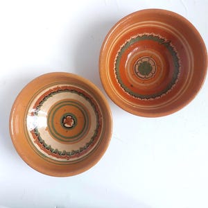 Op de afbeelding: Twee terracotta keramische kommen met ingewikkelde, handgeschilderde ontwerpen. De kommen hebben concentrische cirkels in oranje, groen en wit, met een bloemmotief in het midden. De randen zijn een donkerdere tint oranje.
