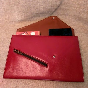 May include: A red leather clutch with a zippered pocket and a brown flap. The flap has a small metal button closure. The clutch is open and has a small book and a black wallet inside.