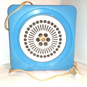 Op de afbeelding: Een vintage blauwe elektrische ventilator met een wit cirkelvormig rooster. De ventilator heeft een vierkant frame met afgeronde hoeken. Een bruin stroomsnoer en stekker zijn bevestigd. Afmetingen: ongeveer 20 cm x 20 cm.