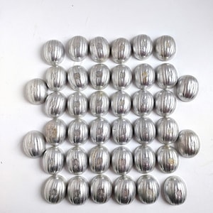 Könnte beinhalten: Eine Sammlung silberfarbener, oval geformter Metallperlen. Jede Perle hat eine gerippte Textur und ein kleines, eingraviertes Design. Die Perlen sind in ordentlichen Reihen vor einem weißen Hintergrund angeordnet, was auf ein Bastel- oder Schmuckthema hindeutet.