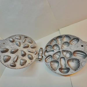 May include: An open, antique, cast-iron waffle iron with a hinged design. One side features a pattern of various-sized stones, while the other side has seashell shapes. The metal has a weathered, silver finish, suggesting age and use.
