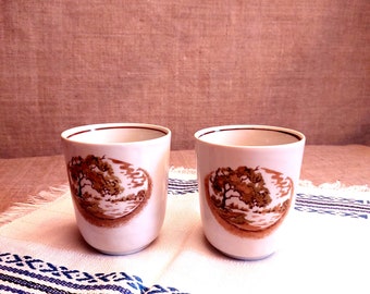 以下が含まれることがあります： 茶色と金の風景デザインが施された、2つの白いセラミックカップ。カップは丸みを帯びた形状で、茶色の縁があります。白と青の模様の布の上に置かれています。
