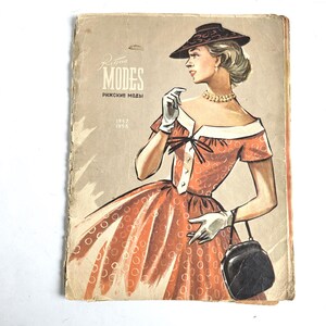 May include: A vintage 1957-1958 fashion magazine cover titled Rīgas MODES featuring an illustration of a person wearing an orange dress with a white circle pattern, a dark hat, white gloves, a beaded necklace, and carrying a black handbag.
