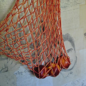 May include: A red and yellow string mesh bag filled with red apples. The bag is hanging on a wall with a newspaper and a drawing of a deer.