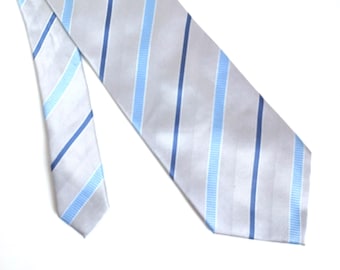 May include: A light gray tie with vertical stripes in varying shades of blue. The tie is made of a smooth, shiny fabric and is angled across a white background. The stripes are evenly spaced and create a classic, professional look.