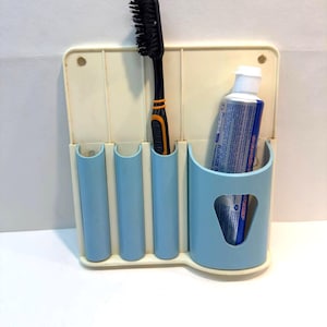 May include: A vintage toothbrush holder in off-white and light blue. It features three small, curved compartments and a larger one for a tube of toothpaste. A black toothbrush with orange accents is in one slot.