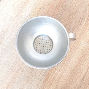 Könnte beinhalten: Ein silberfarbener Metalltrichter mit einem perforierten Sieb in der Mitte. Der Trichter hat eine breite, schalenartige Form mit einem kleinen Griff an der Seite. Der Hintergrund ist eine hellbraune Holzoberfläche.