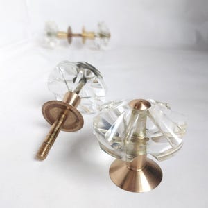May include: Two clear glass cabinet knobs with a faceted design and gold metal bases. The knobs have a screw-on design.