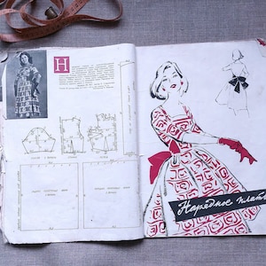 May include: Open vintage sewing pattern book featuring dress illustrations and diagrams. The book shows a red and white patterned dress design, with the text "Нарядное платье" on the cover. A measuring tape and thimble are visible.