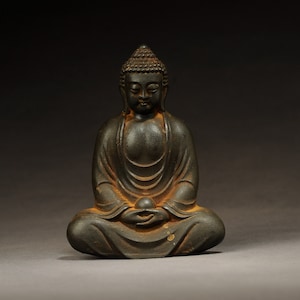 Op de afbeelding: Een donker bronzen beeld van een zittende Boeddha met een serene uitdrukking. Het beeld is gedetailleerd met ingewikkelde houtsnijwerk en heeft een verweerde patina.