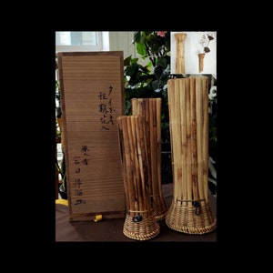 May include: Three brown bamboo vases with a woven base. The vases are different sizes and are arranged in a row. The largest vase is on the right and the smallest vase is on the left. The vases are sitting on a brown surface. The text on the wooden box behind the vases reads "花入れ" in Japanese.