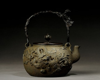 May include: A dark gray cast iron teapot with a dragon and floral design. The teapot has a black wooden handle and a round knob on the lid.
