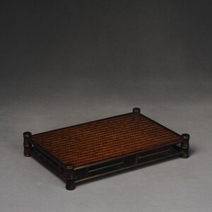 Puede incluir: Una bandeja rectangular de bamb&uacute; tejido marr&oacute;n oscuro con un marco lacado en negro y cuatro patas. La bandeja es rectangular y tiene un borde elevado.