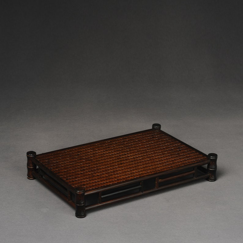 Puede incluir: Una bandeja rectangular de bamb&uacute; tejido marr&oacute;n oscuro con un marco lacado en negro y cuatro patas. La bandeja es rectangular y tiene un borde elevado.