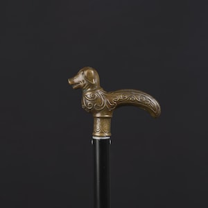 May include: A walking stick featuring a metallic bronze-toned handle shaped like a dog head with intricate swirling scrollwork patterns. The handle is attached to a smooth, straight black shaft, creating a contrast between the ornate metal top and the simple dark base.