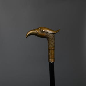 May include: A bronze-colored walking cane with an eagle head handle. The eagle head is detailed with feathers and a curved beak. The cane's shaft is black, and the handle has a decorative band.