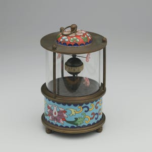 May include: A small, round, brass clock with a glass dome. The clock face is visible through the glass and has black Roman numerals. The clock is decorated with colorful enamel flowers and swirls.